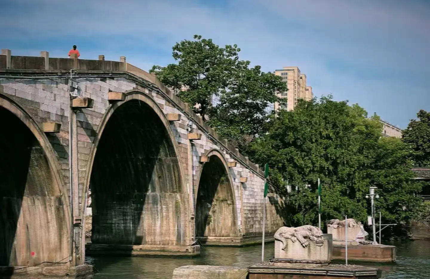 Stone arched bridge over a river with green trees nearby; a person in an orange shirt stands on the walkway above.