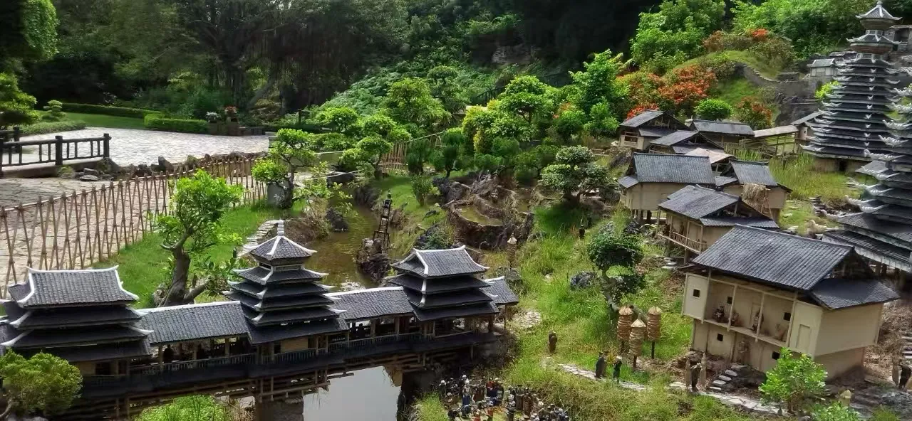 Miniature traditional Asian village with tiered pagoda buildings, a wooden bridge, and lush garden scenery.