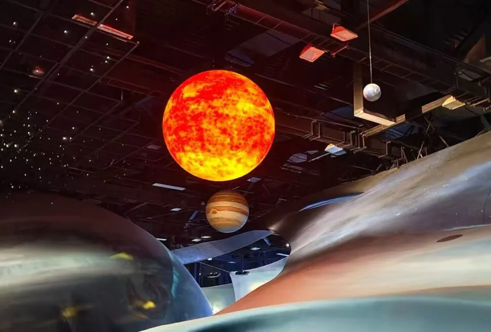 Large bright orange sun model suspended from the ceiling with a Saturn-like planet nearby in a dark museum exhibit.