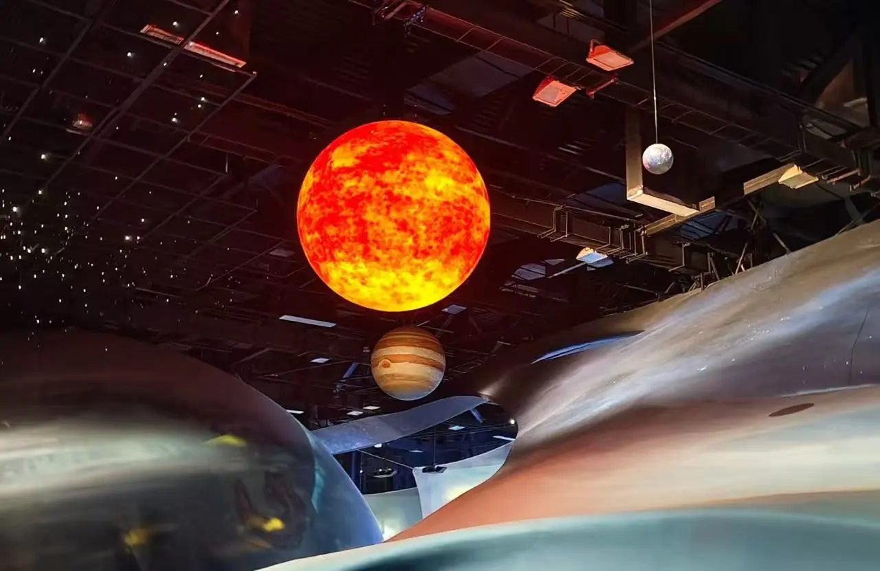 Guangzhou Science Center,Guangzhou,Chinatour Large bright orange sun model suspended from the ceiling with a Saturn-like planet nearby in a dark museum exhibit.
