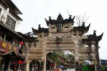 Ornate stone Chinese paifang arch over a bustling street lined with shops and red lanterns.