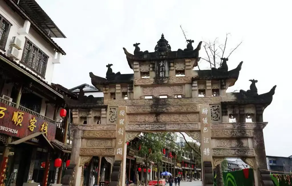 Jianmen Pass Ancient Town,Mianyang,Chinatour Ornate stone Chinese paifang arch over a bustling street lined with shops and red lanterns.