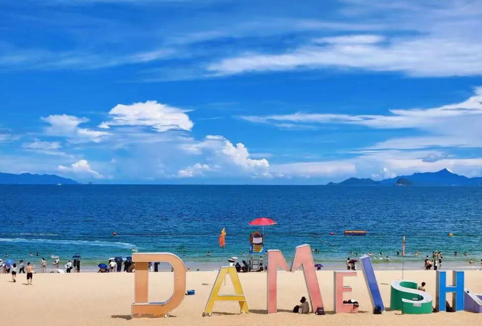 Oversized colorful letters spelling a word on a sandy beach with the blue sea and people in the background.