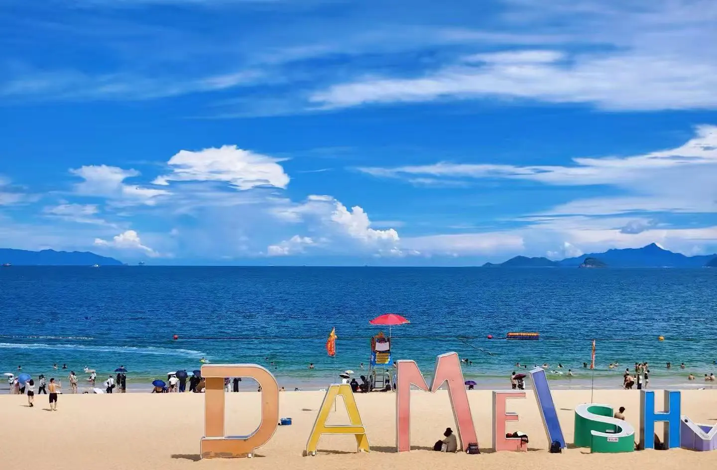 Oversized colorful letters spelling a word on a sandy beach with the blue sea and people in the background.