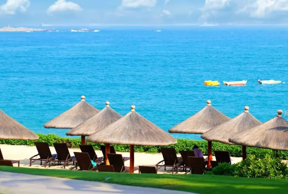 Yalong Bay National Resort Area,Sanya,Chinatour Tropical beach with thatched umbrellas and lounge chairs facing a blue sea; small boats drift on calm water.