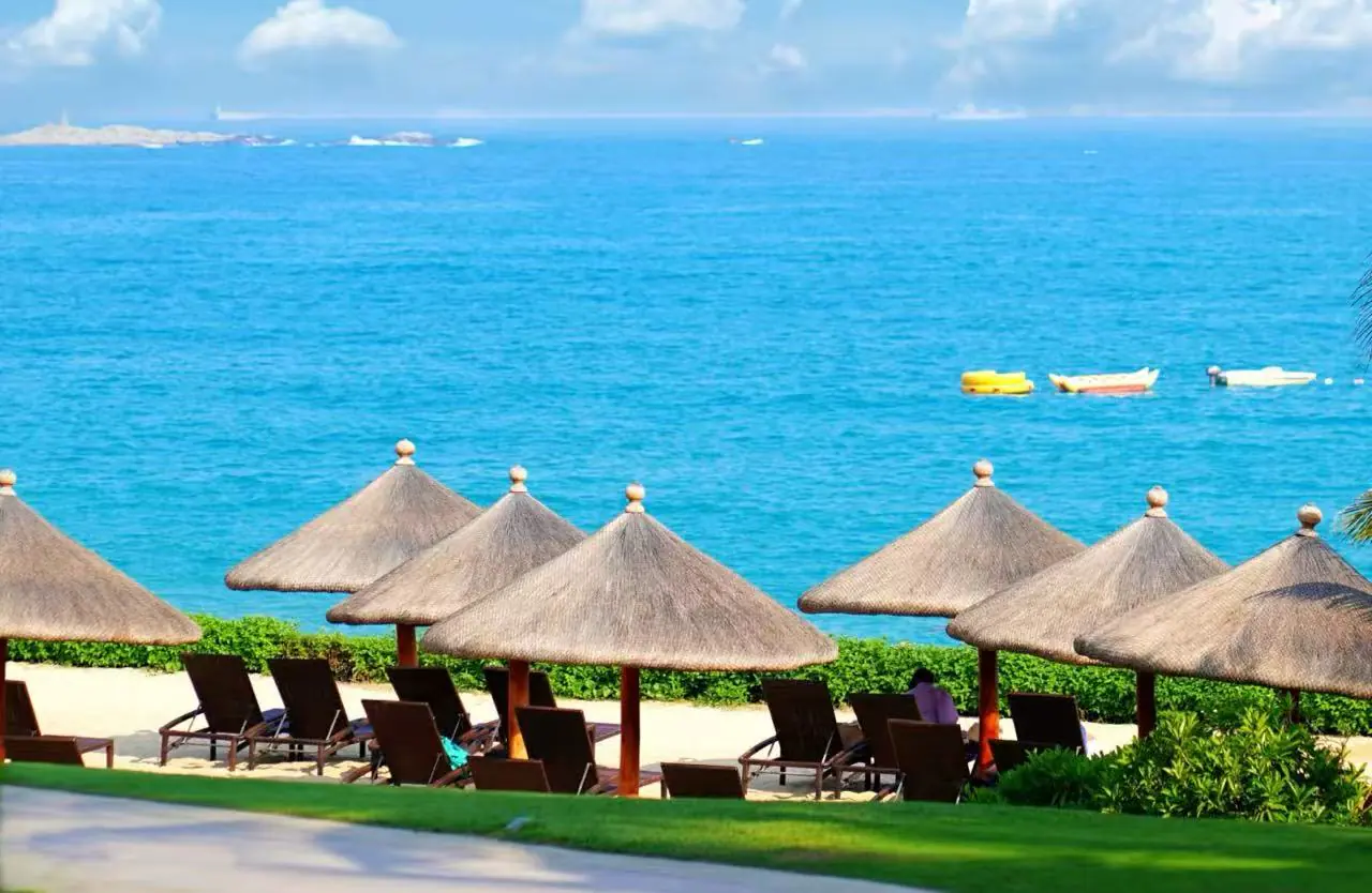 Yalong Bay National Resort Area,Sanya,Chinatour Tropical beach with thatched umbrellas and lounge chairs facing a blue sea; small boats drift on calm water.