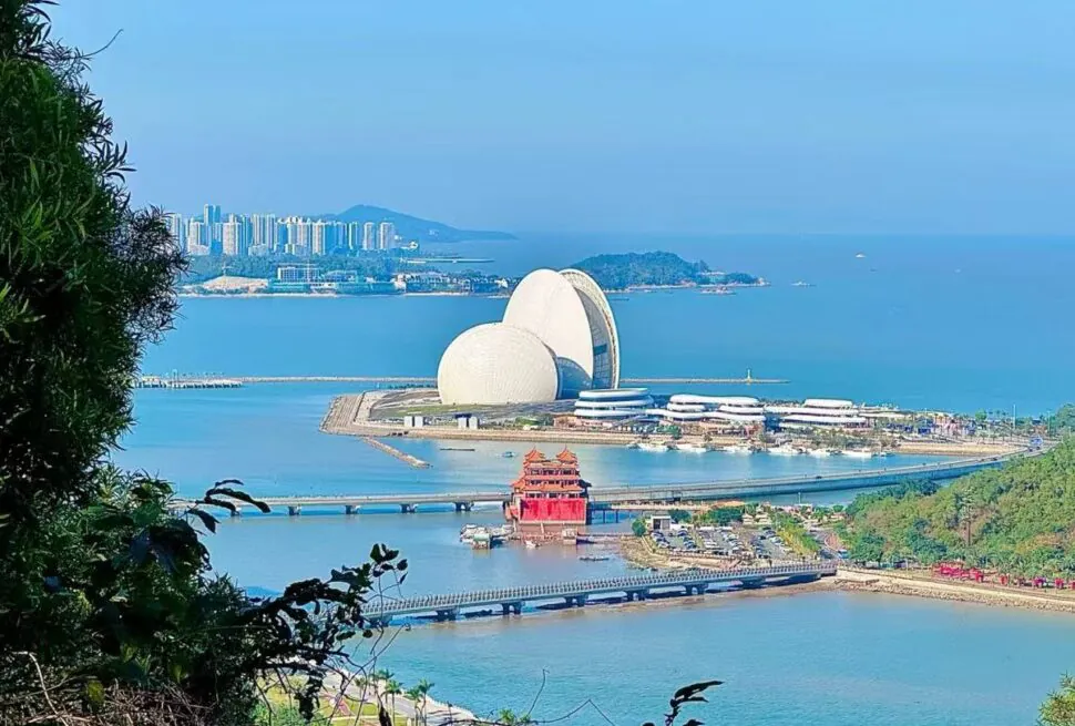 Coastal panorama with white futuristic domes on a peninsula, a red pagoda and a bridge over blue water.