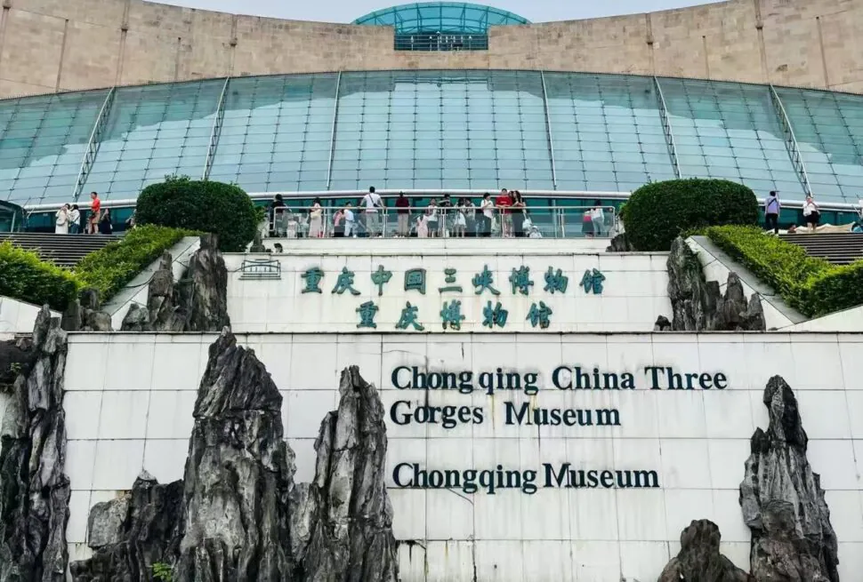 Exterior view of Chongqing China Three Gorges Museum with a glass-domed atrium, visitors on the terrace, and rock sculptures framing a sign that reads 'Chongqing China Three Gorges Museum' in English and Chinese.