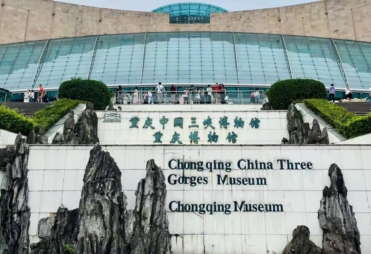 Chongqing Three Gorges Museum,Chongqing,Chinatour Exterior view of Chongqing China Three Gorges Museum with a glass-domed atrium, visitors on the terrace, and rock sculptures framing a sign that reads 'Chongqing China Three Gorges Museum' in English and Chinese.
