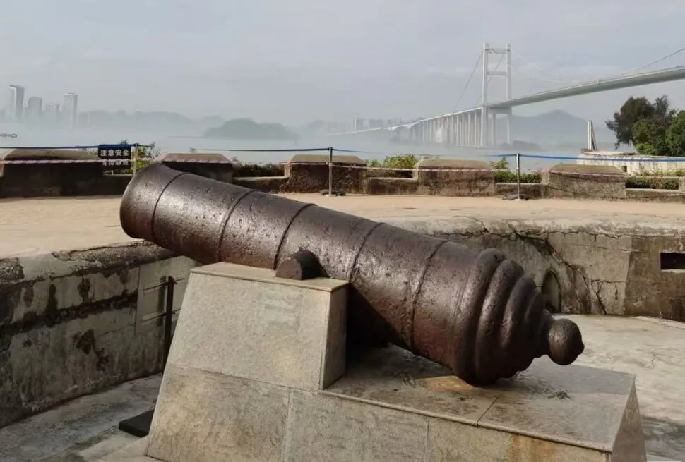 Humen Fort,Dongguan,Chinatour Antique rusted cannon mounted on a stone pedestal at a coastal fort, with a misty city skyline and a suspension bridge in the background.