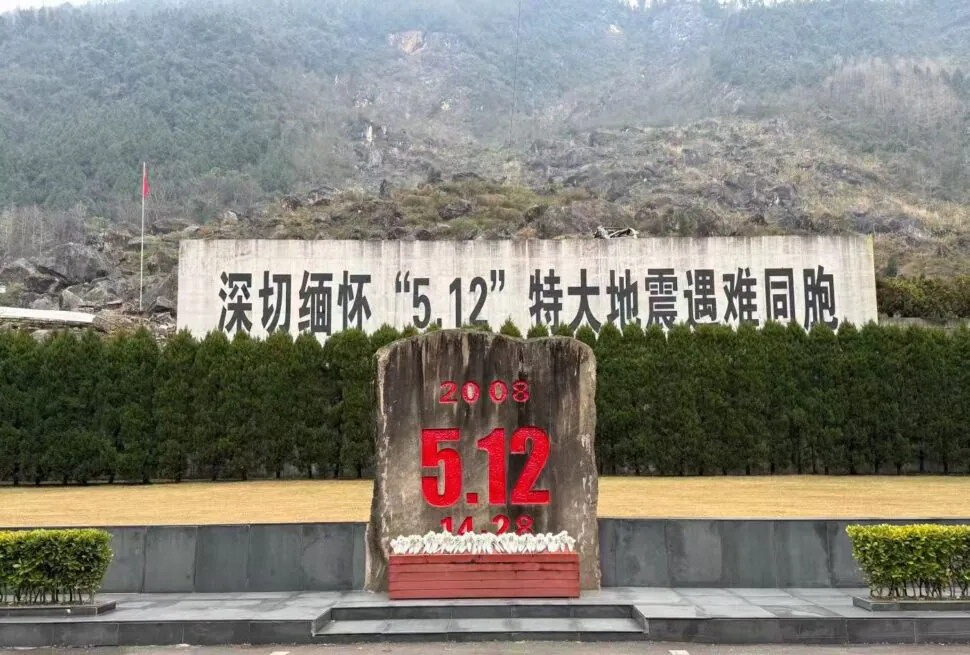 Monument with a large red '5.12' and '2008' on a rock in front of a hedge and Chinese text backdrop, mountains in the background (Earthquake memorial).