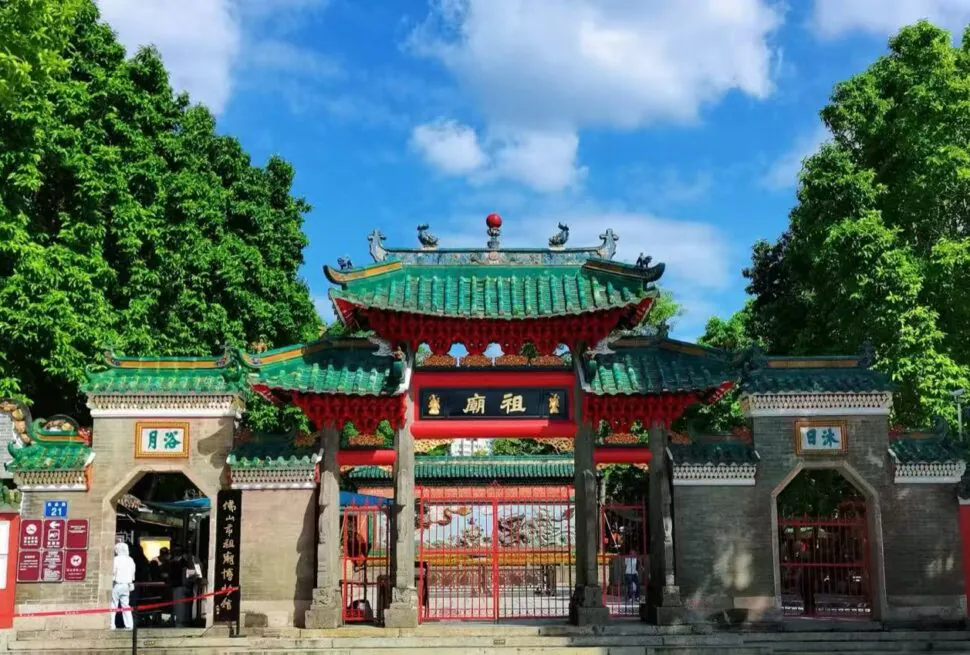 Ancestral Temple ,Guangzhou,Chinatour Traditional Chinese temple gate with a green tiled roof, red pillars, and ornate Details, set between lush trees under a blue sky.