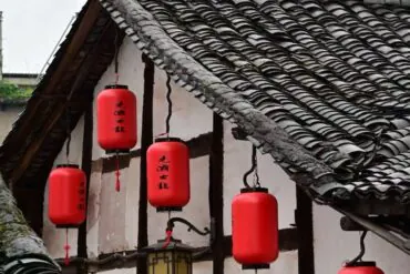 Red cylindrical Chinese lanterns hanging from a traditional tiled roof with white walls and wooden beams, daytime scene