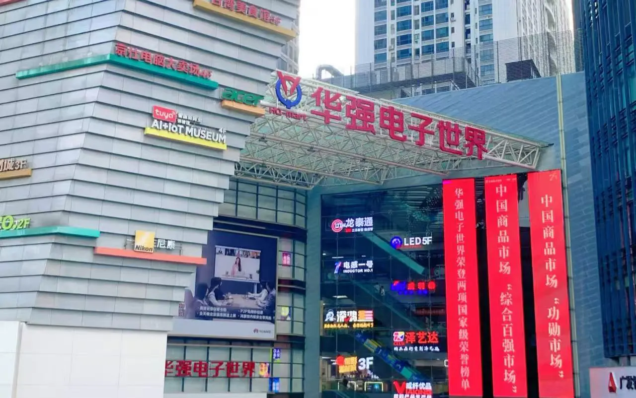 Exterior view of the Huaqiang Electronic World shopping complex with a glass facade, red vertical banners, and colorful brand signs on the building exterior, in a busy urban area.