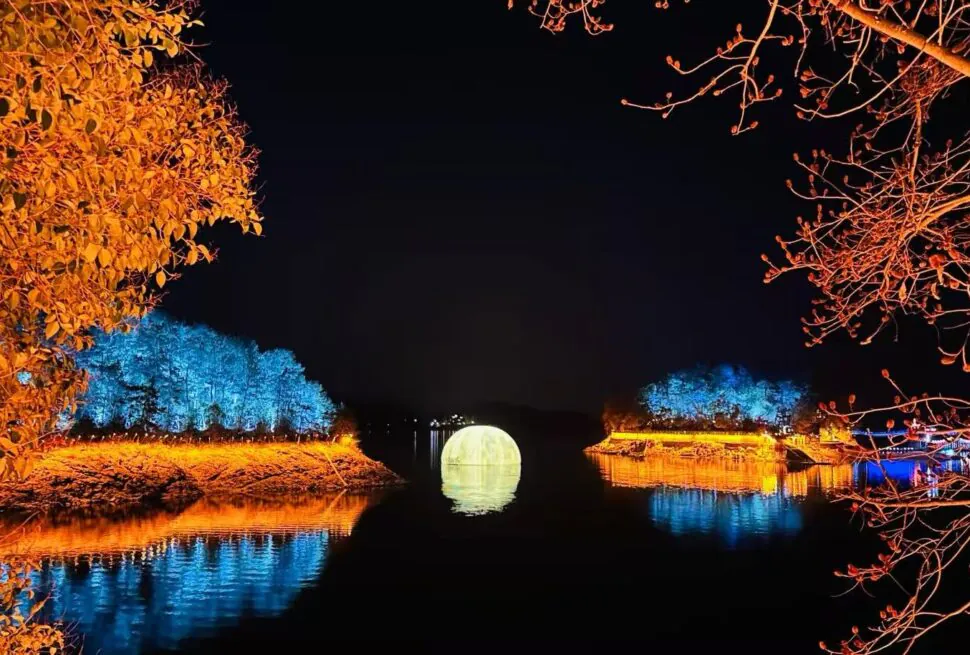 Night scene of a round illuminated bridge over a calm river, with orange-lit trees along the banks and blue-lit trees in the background reflecting in the water, at night.