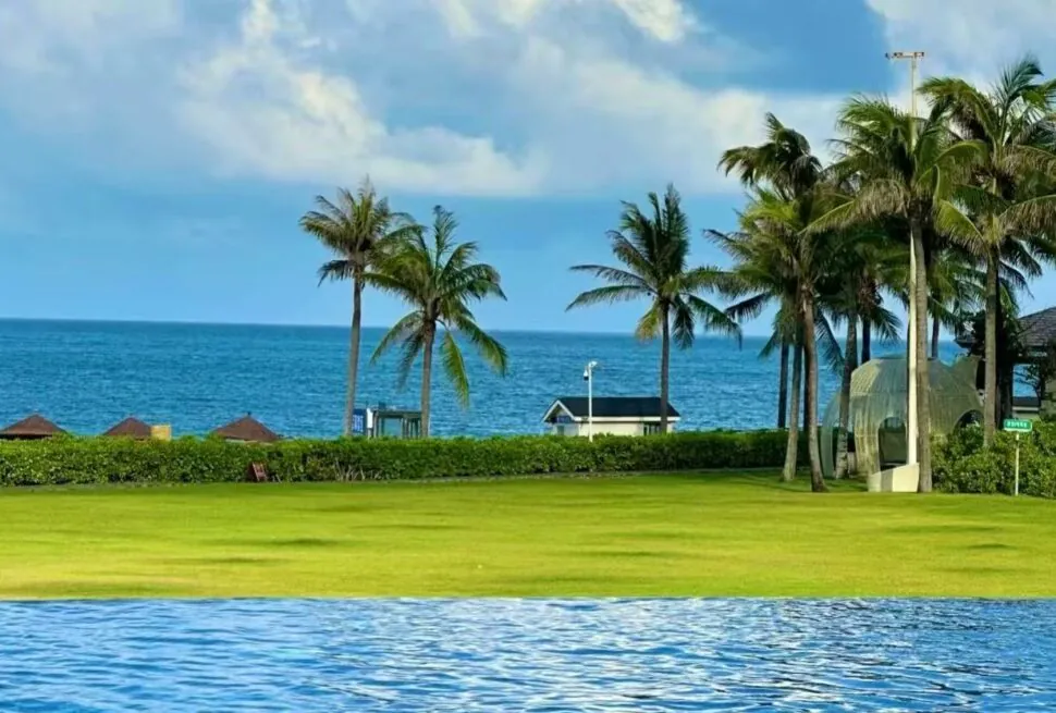 Sanya Bay Coastal ,Sanya,Chinatour Poolside view at a tropical resort with a green lawn, palm trees, and a blue ocean beyond the hedges.