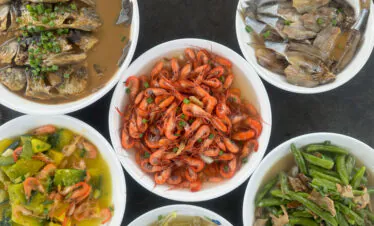Assortment of cooked seafood dishes in white bowls: vibrant shrimp in the center with fish pieces and mixed vegetables around it.