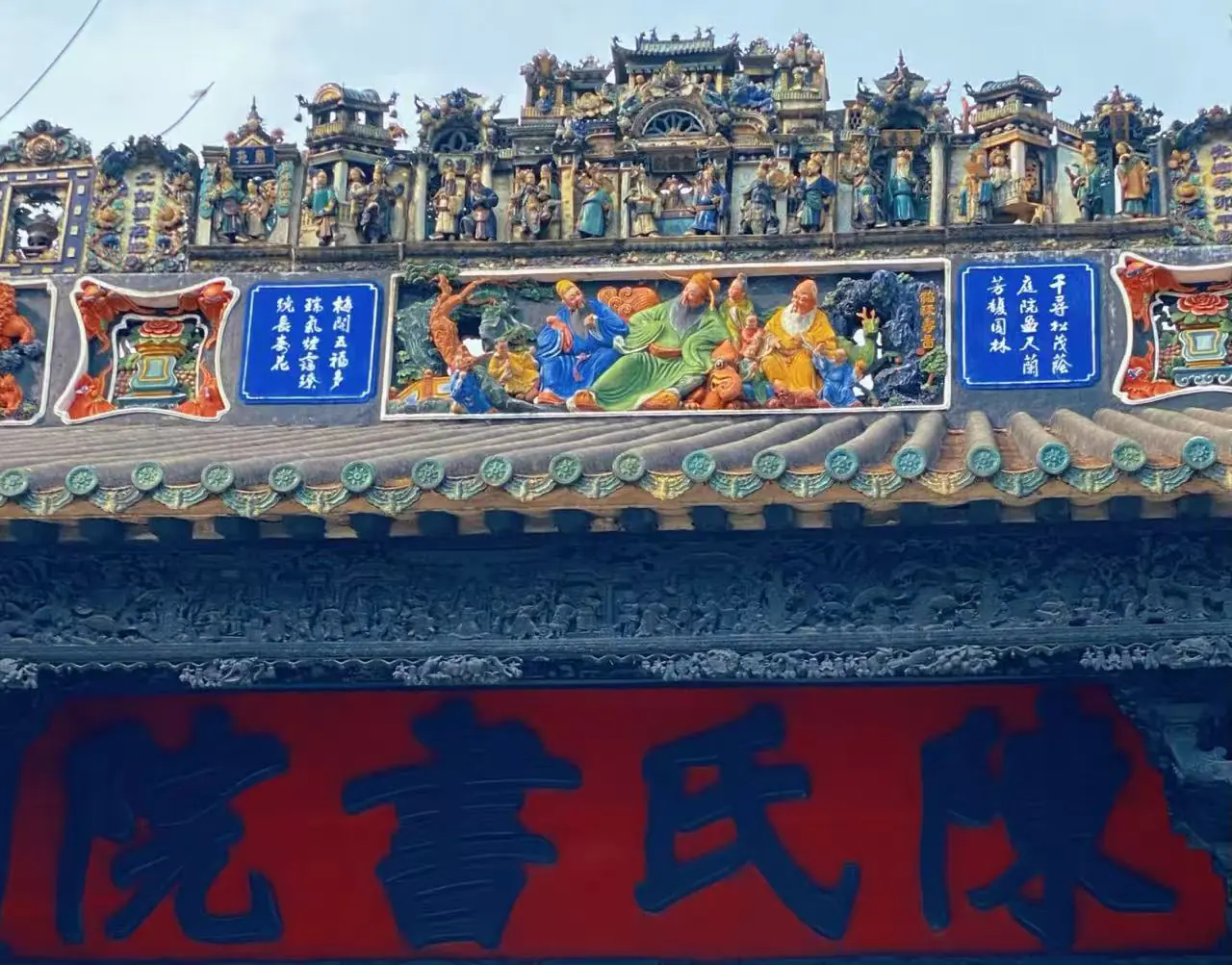 Chen Clan Ancestral Hall,Guangzhou,Chinatour Ornate temple roof with colorful carvings and figural sculptures, blue plaques with Chinese characters, and a large red banner beneath.