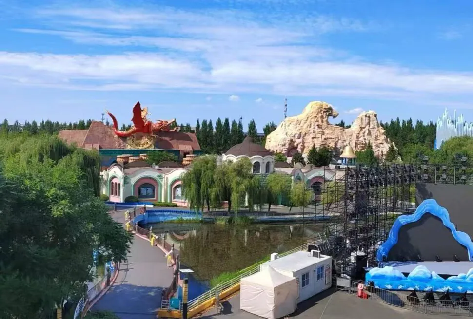 Changchun Movie Wonderland,Changchun,Chinatour A colorful theme park with a red dragon statue atop buildings, green trees, and rocky sculptures in the background under a blue sky.
