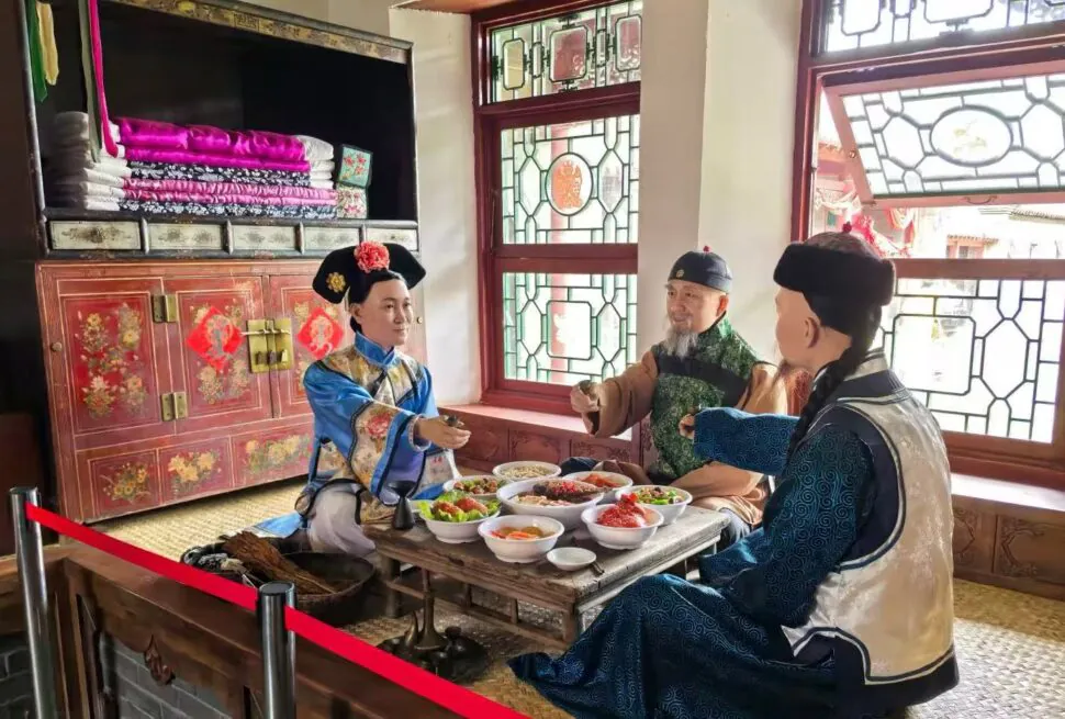 Three people in traditional Chinese imperial attire share a low table meal in a decorated room with bowls of food and red lattice windows behind them.