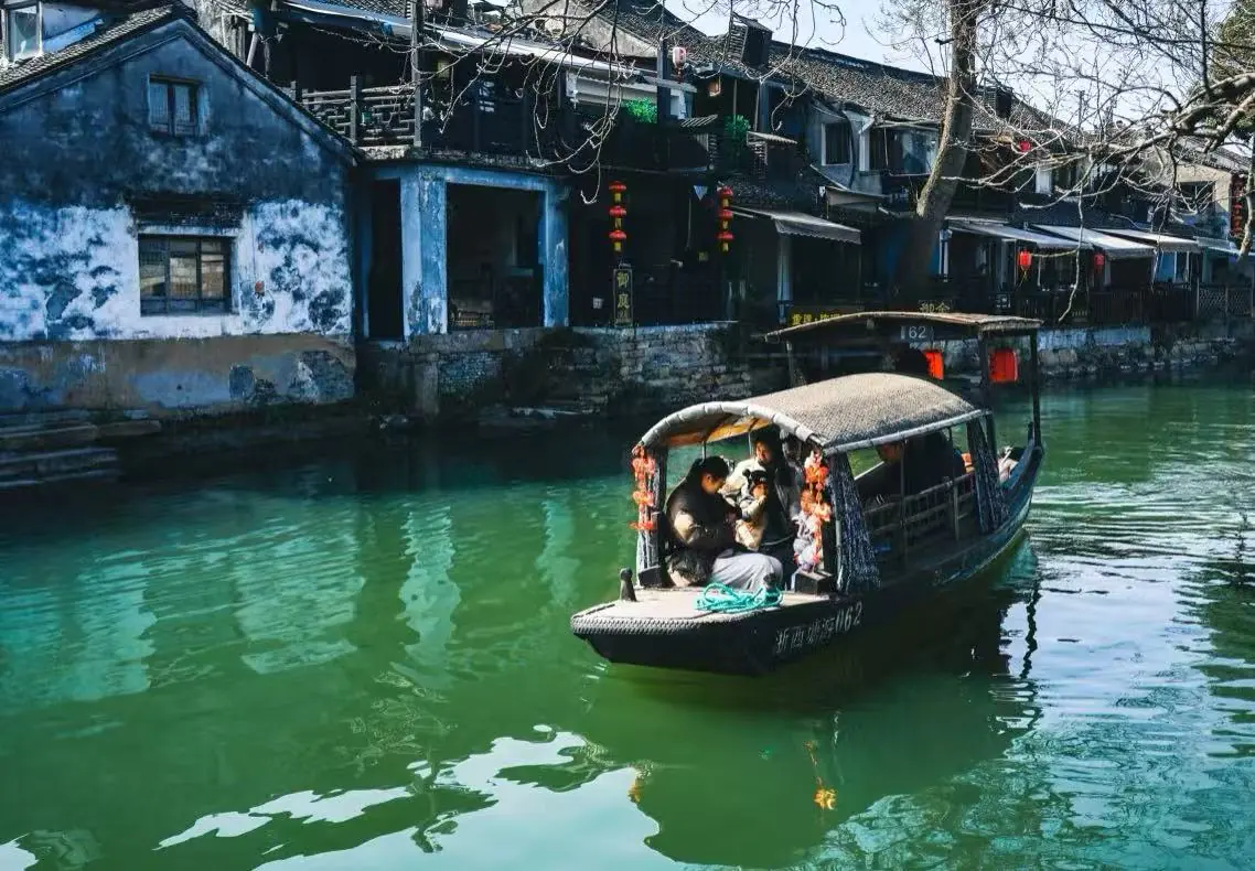 Xitang Water Town,Hangzhou,Chinatour Tour boat with a woven canopy glides along a green canal beside blue-washed houses adorned with red lanterns. Family passengers sit inside the boat.