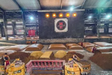 Interior of a large warehouse with sacks and piles of grain or powder, workers in red vests processing materials near wooden barrels and a central stage with Chinese decor.