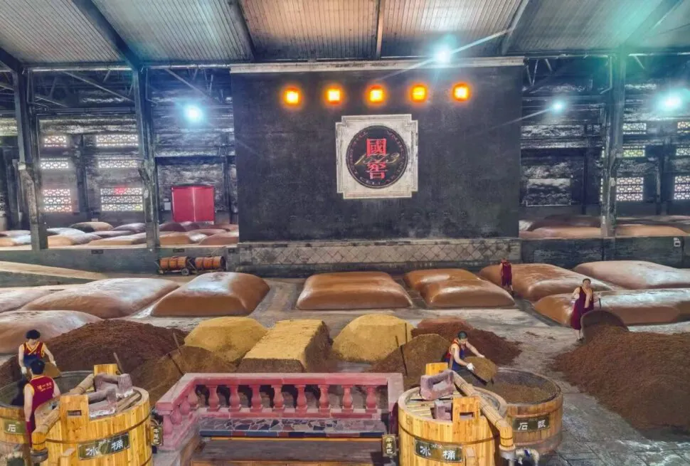 Interior of a large warehouse with sacks and piles of grain or powder, workers in red vests processing materials near wooden barrels and a central stage with Chinese decor.