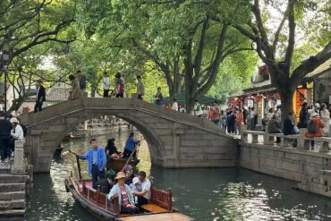 Stone arched pedestrian bridge over a canal with people walking; boats on the water and trees overhead on a sunny day.