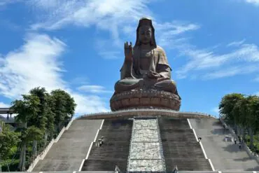 Giant seated statue of Guanyin atop a wide stone staircase under a blue sky.