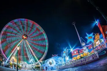 Nighttime carnival scene with a brightly lit spinning Ferris wheel and colorful rides along a illuminated promenade at a fairground edge nearby edges of the park.