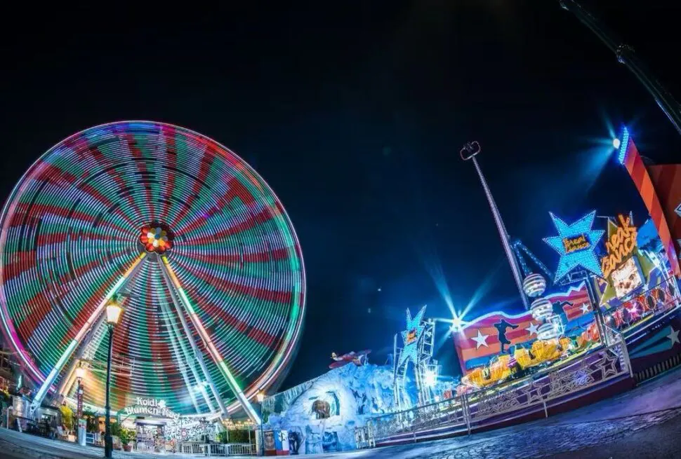 Nighttime carnival scene with a brightly lit spinning Ferris wheel and colorful rides along a illuminated promenade at a fairground edge nearby edges of the park.