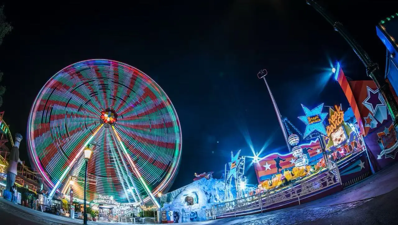 Nighttime carnival scene with a brightly lit spinning Ferris wheel and colorful rides along a illuminated promenade at a fairground edge nearby edges of the park.