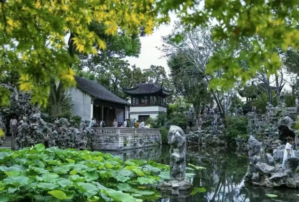 People walk along a stone terrace beside a pond in a traditional Chinese garden with rock formations and a pagoda-style pavilion.
