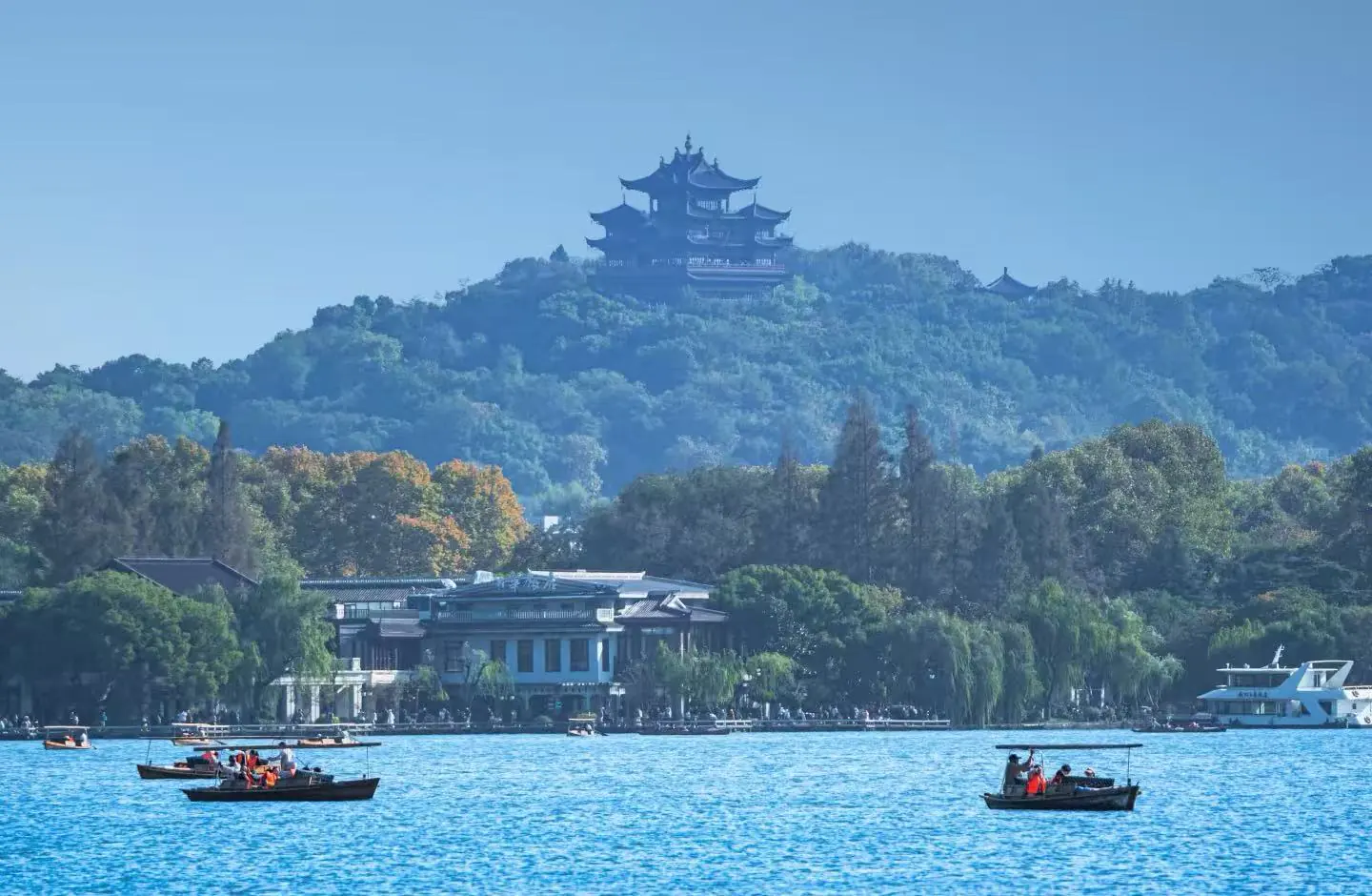 West Lake,Hangzhou,Chinatour Boats carry passengers on a blue lake with a tree-covered hill and a distant multi-tier pagoda.