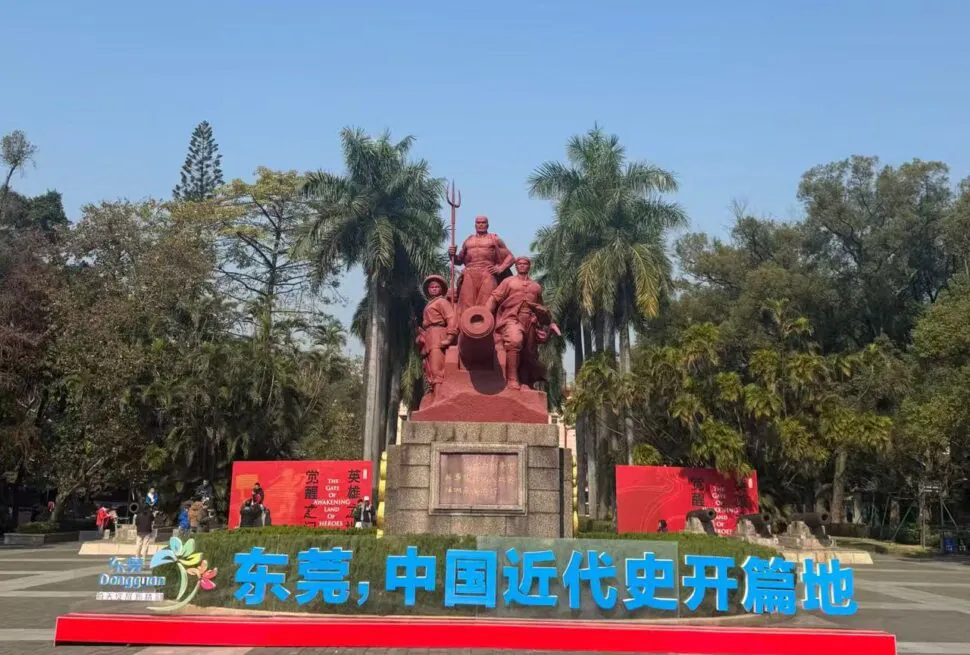Guangdong Opium War Museum,Dongguan,Chinatour Red statues group with a cannon on a stone pedestal in a park, palm trees behind, blue sky above, bold blue Chinese lettering in front.
