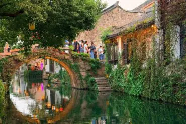 Stone arch bridge over a green canal, ivy-covered walls, and people strolling along a historic riverside walkway.
