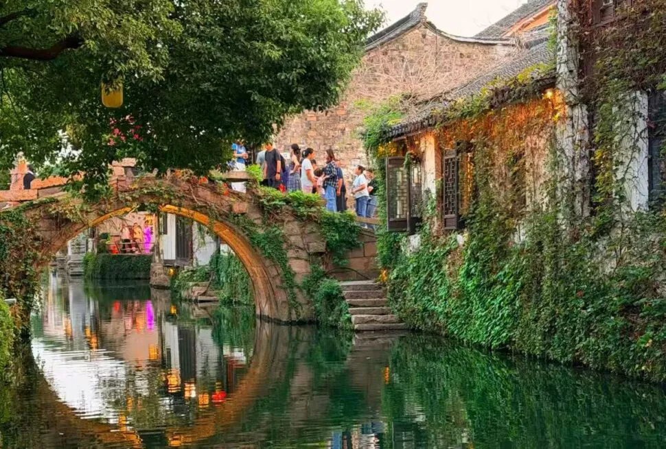 Stone arch bridge over a green canal, ivy-covered walls, and people strolling along a historic riverside walkway.
