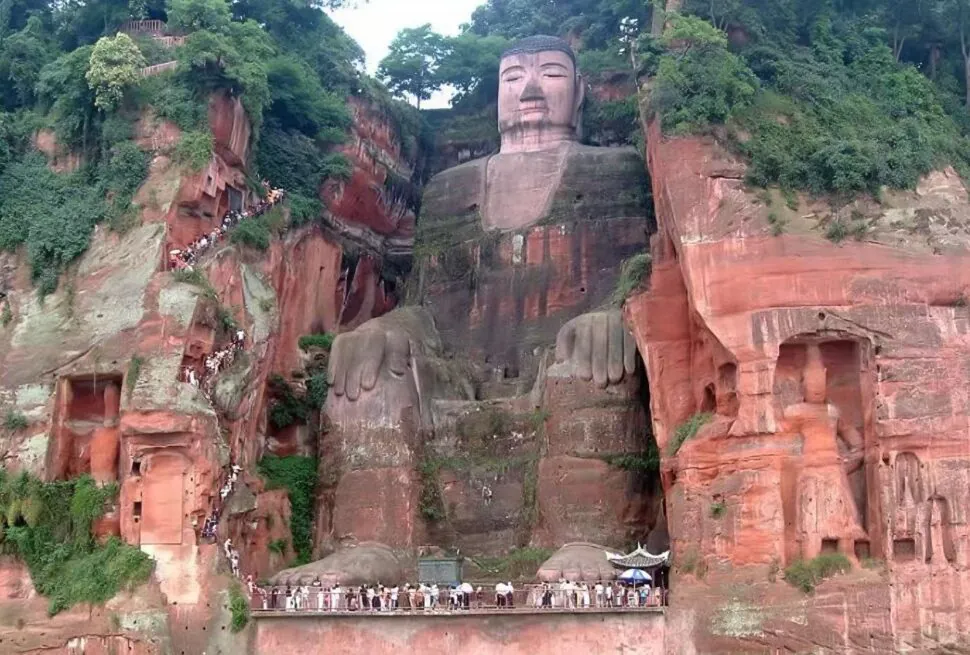 Giant Buddha statue carved into a red cliff with a calm face and large hands, surrounded by greenery and visitors along a stair path on the left.