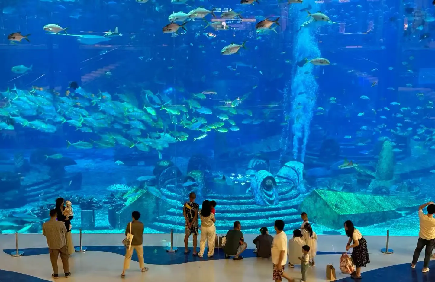 Sanya Atlantis Waterpark,Sanya,Chinatour Visitors observe a large blue aquarium tank filled with a school of fish and a decorative sunken ship centerpiece behind the glass.