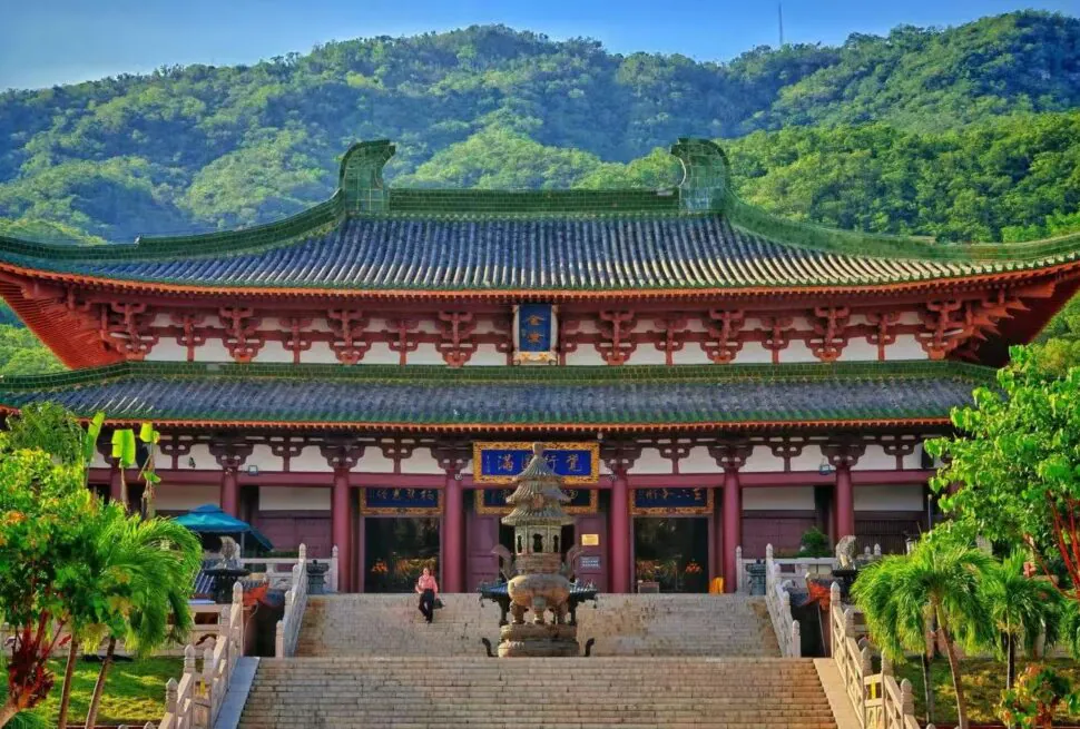 Temple with red and green tiled roof, stone stairs, and a bronze incense burner, set against a forested hillside.