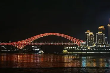 Nighttime view of a red-lit arched bridge spanning a river with a city skyline to the right; reflections glow on the water.