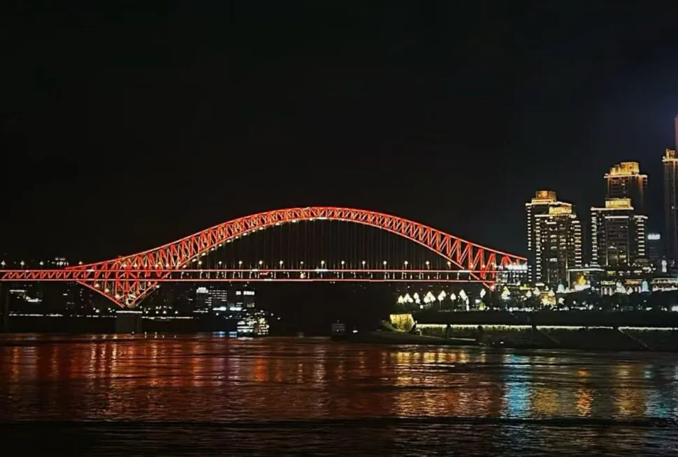 Nighttime view of a red-lit arched bridge spanning a river with a city skyline to the right; reflections glow on the water.