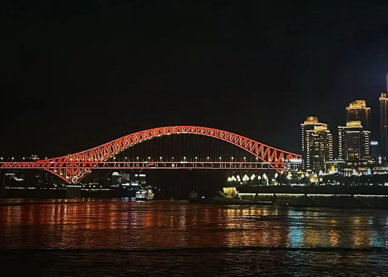 Chaotianmen Dock and Riverside Promenade,Chongqing,Chinatour Nighttime view of a red-lit arched bridge spanning a river with a city skyline to the right; reflections glow on the water.