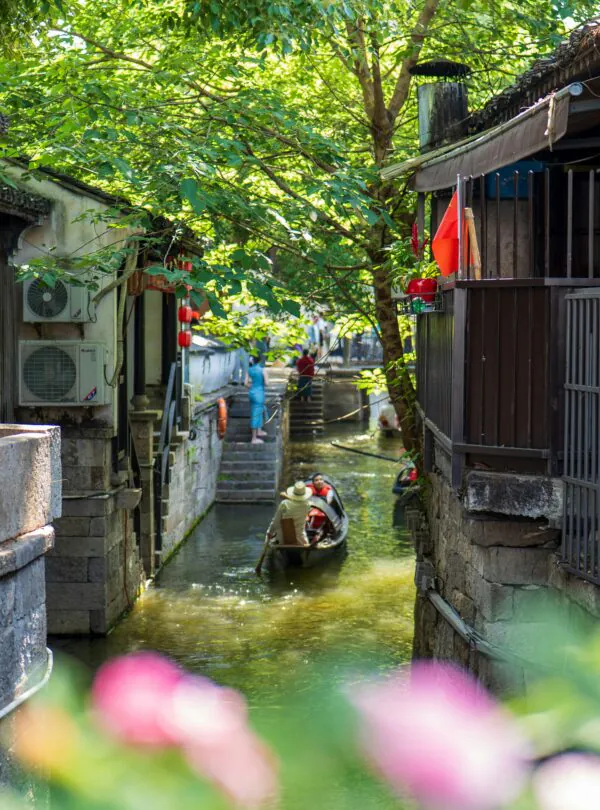 A narrow canal between traditional buildings with a small boat and people paddling, surrounded by green trees.