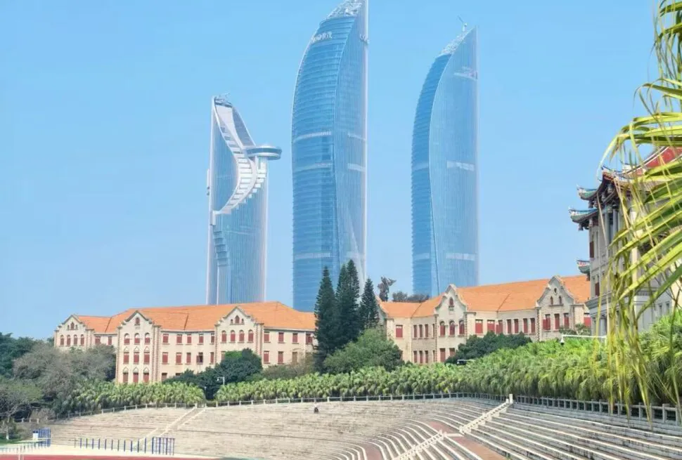 Beige campus buildings with red roofs sit before a skyline of three blue glass skyscrapers; a sports field and empty bleachers in the foreground.