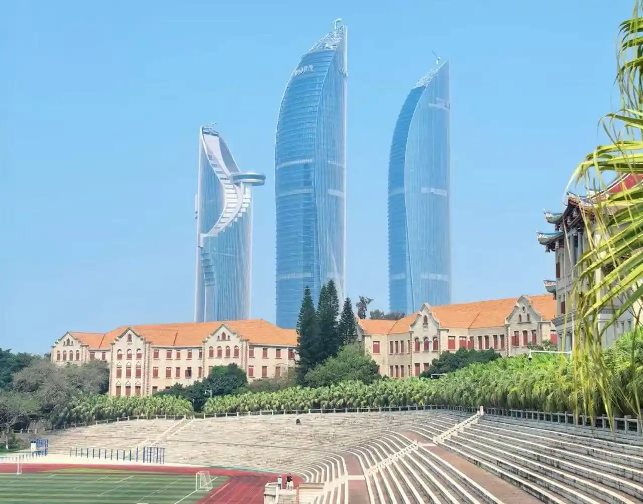 Xiamen University,Xiamen,Chinatour Beige campus buildings with red roofs sit before a skyline of three blue glass skyscrapers; a sports field and empty bleachers in the foreground.