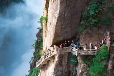 Visitors wearing orange helmets walk along a railinged cliff path carved into the rocky mountainside beside a misty waterfall to the left.