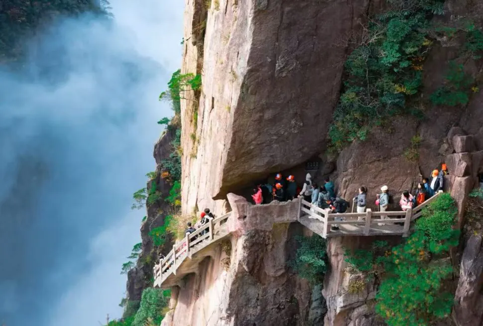Visitors wearing orange helmets walk along a railinged cliff path carved into the rocky mountainside beside a misty waterfall to the left.