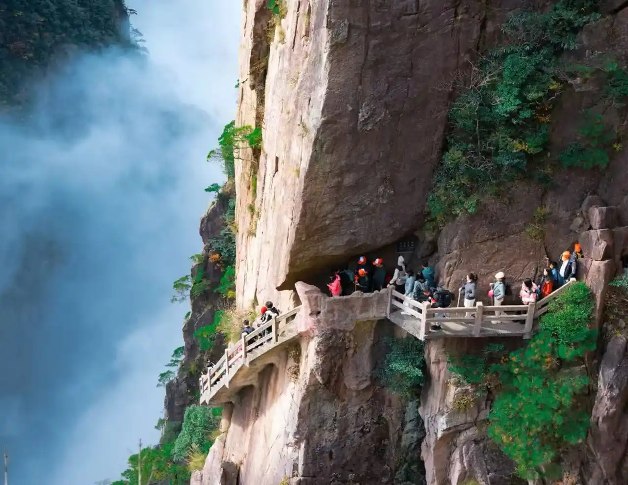 Xihai Grand Canyon,Huangshan,Chinatour Visitors wearing orange helmets walk along a railinged cliff path carved into the rocky mountainside beside a misty waterfall to the left.