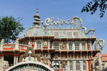 Elaborately decorated mosaic building with curved, organic shapes, stone lattice windows, and a large cursive sign on the roof against a blue sky.